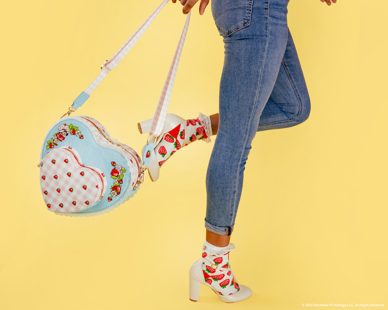 Woman wearing skinny blue jeans with strawberry socks and white heels, with one leg bent, and swinging the pink and blue Strawberry Shortcake denim heart-shaped crossbody bag behind her, standing against a yellow background.